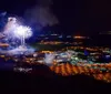 The image captures a nighttime aerial view of a town brightly illuminated with glowing streets and buildings while fireworks erupt in the sky above