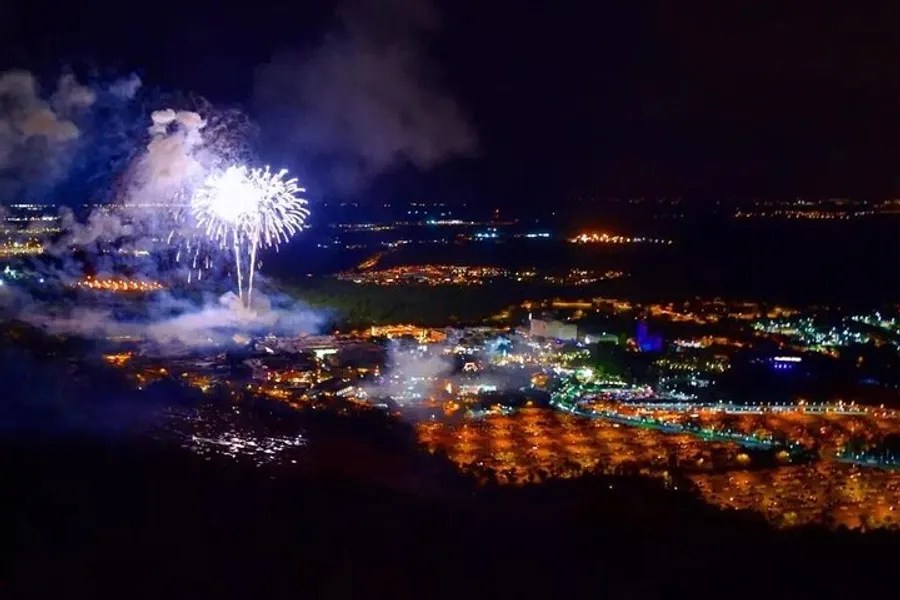 The image captures a nighttime aerial view of a town brightly illuminated with glowing streets and buildings while fireworks erupt in the sky above.