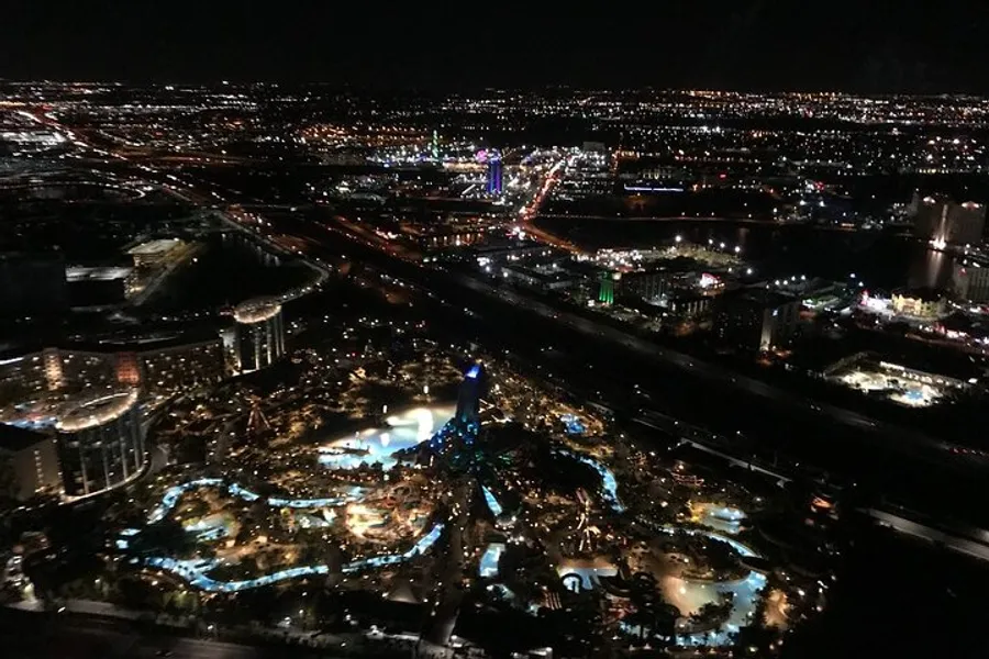 The image is an aerial night view of a brightly lit urban area with distinctive blue-lit pathways winding through a complex of buildings and attractions.