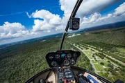The image shows the cockpit view from inside a helicopter flying over a landscape with roads and greenery, highlighting the aircraft's control panel and the horizon in the background.