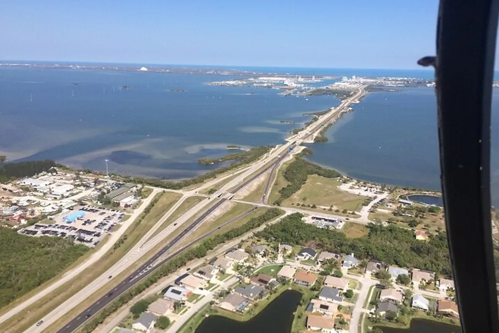The image shows an aerial view of a highway running through a coastal area with residential neighborhoods on one side and a large body of water with docks and boats on the other