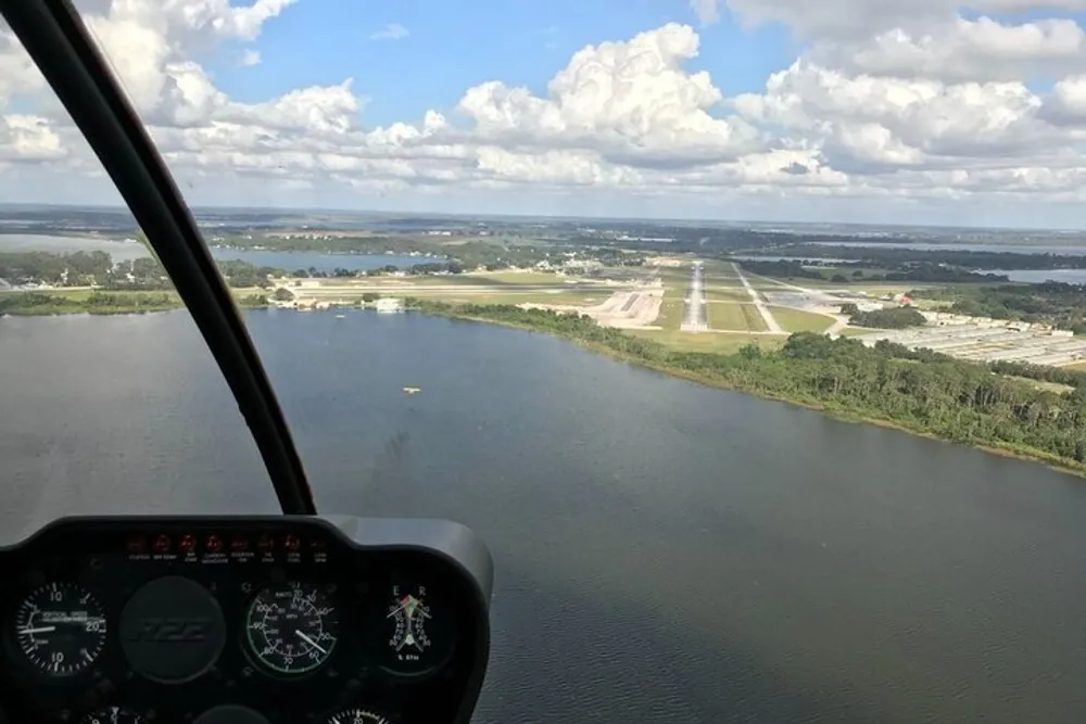 The image shows the cockpit view from a helicopter overlooking an airstrip near a body of water with various buildings and greenery in the distance