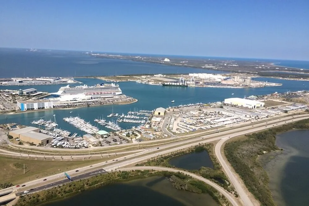The image shows an aerial view of a coastal port area with a cruise ship docked a marina filled with smaller boats several buildings and roads with a vast expanse of water visible in the background