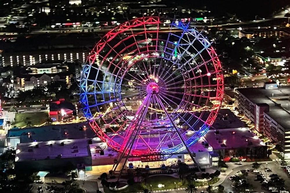 An illuminated Ferris wheel stands out against the night skyline casting vibrant red and blue hues over the surrounding area