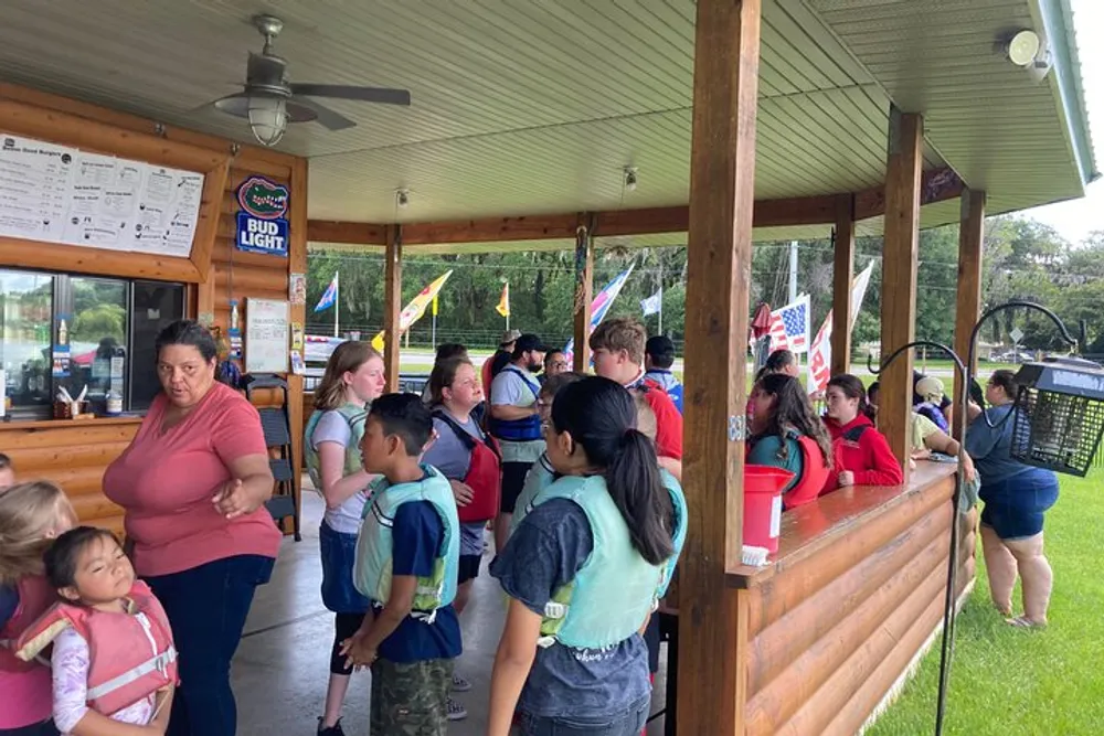A group of people including children are standing in line at an outdoor service counter with some individuals wearing life vests suggesting they might be waiting for a water-related activity
