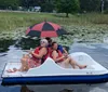 Two green Adirondack chairs and a matching table sit under a red umbrella facing a tranquil lake with a dock and pedal boats