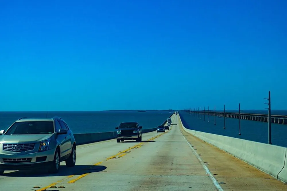 The image shows a sunny day where vehicles are traveling on a long bridge over a calm blue body of water with another parallel bridge visible in the distance