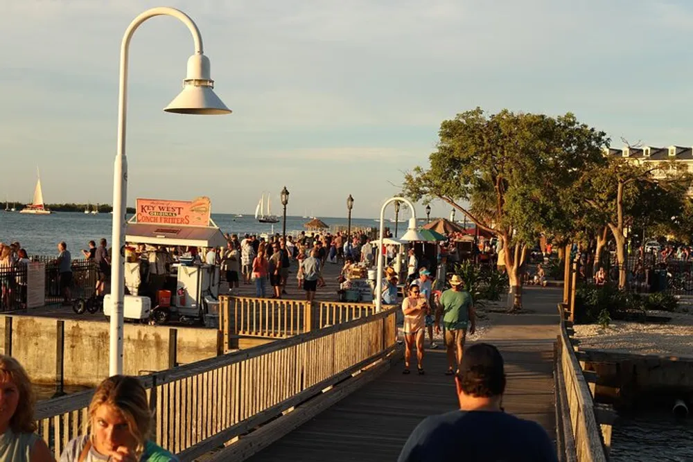 The image shows a bustling boardwalk by the water in Key West with people walking street vendors boats in the distance and a warm sunset ambiance