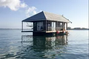 A quaint wooden cabin floats on calm water, featuring a porch with a hanging hammock.