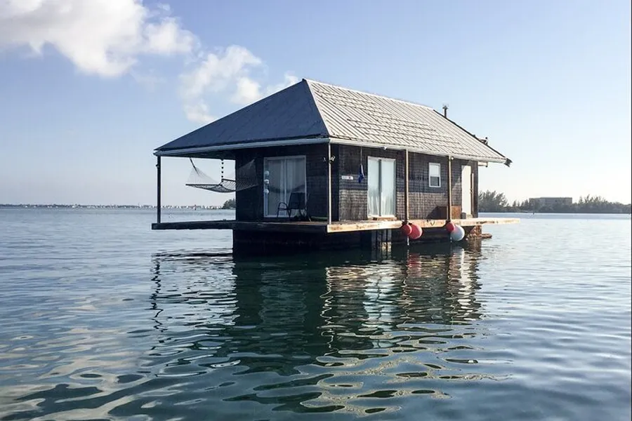A quaint wooden cabin floats on calm water, featuring a porch with a hanging hammock.