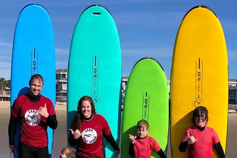 Four individuals in red wetsuits are standing with colorful surfboards on a sunny beach making cheerful hand gestures