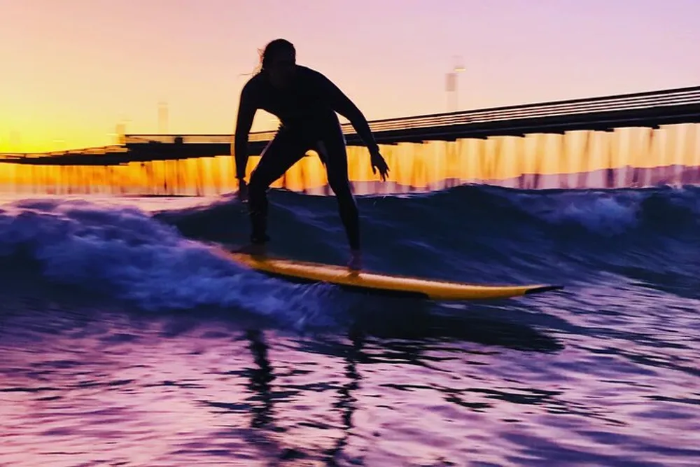 A surfer rides a wave against a backdrop of a colorful sunset and a pier silhouette