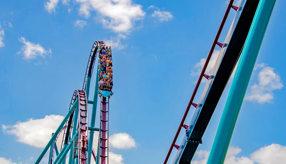 A roller coaster car filled with thrill-seekers is perched at the apex of a steep track ready to plummet against a backdrop of a blue sky scattered with white clouds