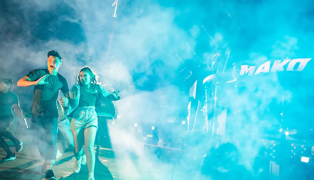 A group of people is running through fog with expressions of excitement with bright lights and the word MAKO visible in the background