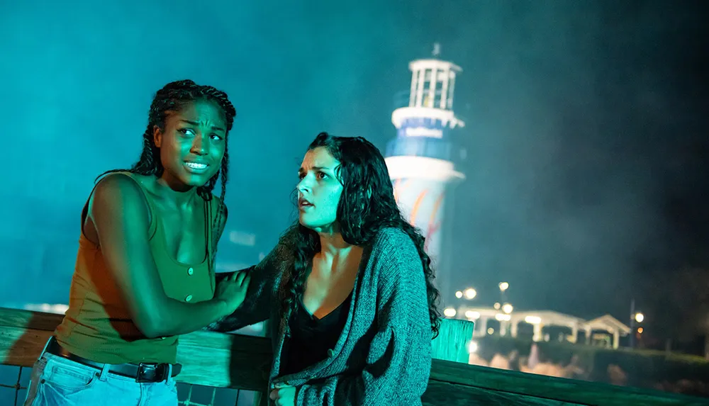 Two women look scared and are clutching each other in front of a misty backdrop with a lighthouse