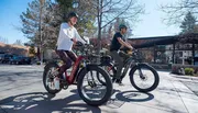 Two people are riding electric bicycles on a sunny day in an outdoor setting with shops in the background.