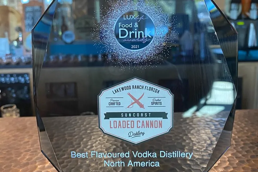 The image shows a clear glass award for 'Best Flavoured Vodka Distillery North America' from 'LUX Life Food & Drink Awards 2021,' sitting on a bar counter, representing an accolade received by the Suncoast Loaded Cannon Distillery based in Lakewood Ranch Florida.