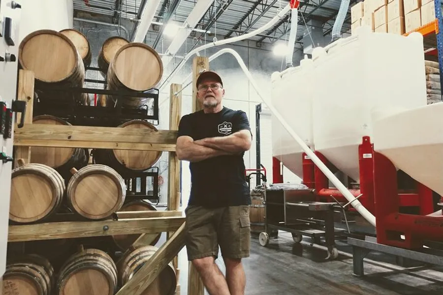 A man is standing with his arms crossed in a brewery or distillery environment, surrounded by wooden barrels and brewing equipment.