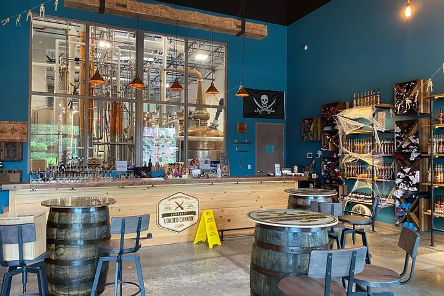 The image shows a spacious distillery tasting room with a distinctive pirate theme, copper distillation equipment visible through a large glass partition, and various merchandise on display.