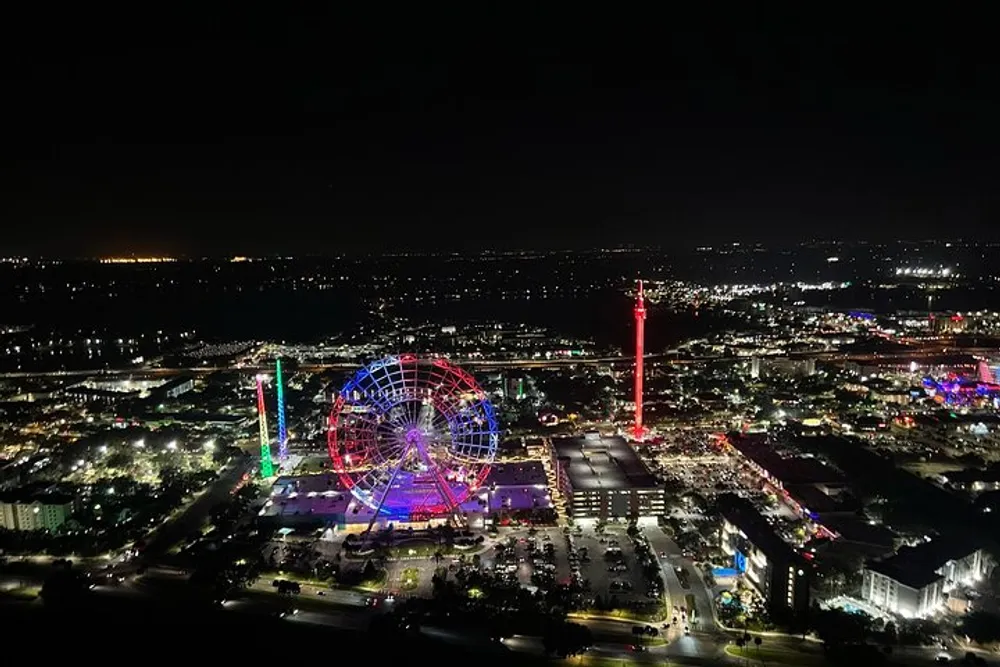 The image shows a vibrant night view of an urban area featuring an illuminated Ferris wheel and other colorful lights from attractions and buildings seen from an elevated perspective