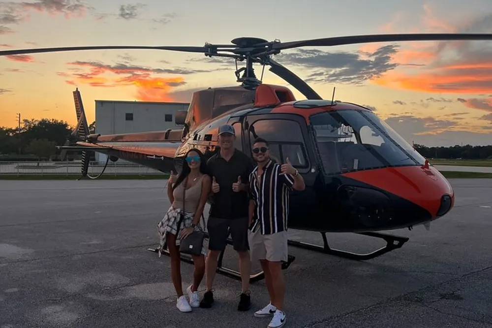 Three people are posing for a photograph with a red and black helicopter in the background and a beautiful sunset sky