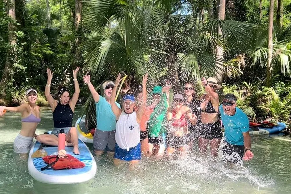 A joyful group of people is having fun in a water setting with some on inflatable tubes and others standing in the water splashing and raising their arms in excitement amid lush greenery