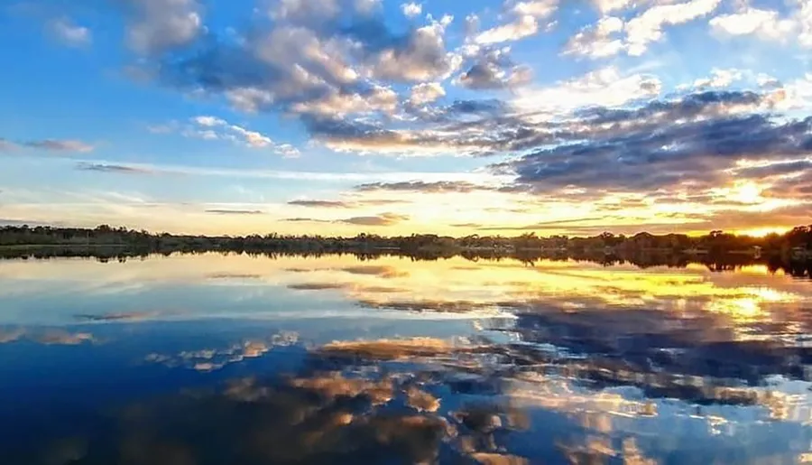 The image shows a dramatic sunset with a sky full of clouds reflected on the calm surface of a large body of water, suggesting a tranquil evening scene.