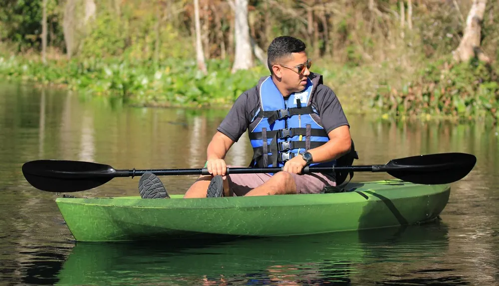 A person is paddling a green kayak on calm water with lush greenery in the background