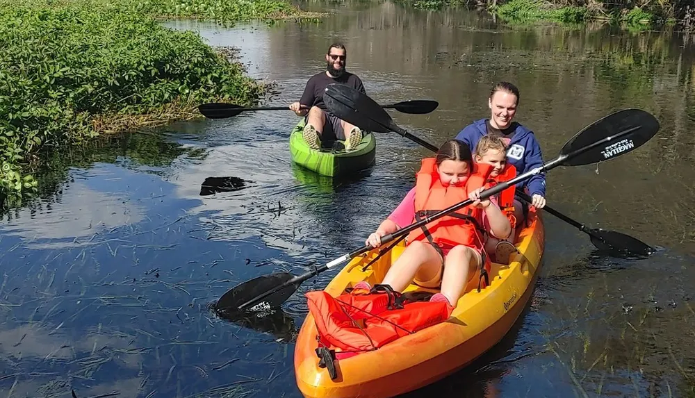 A family is enjoying a kayaking trip on a calm river with a man solo in a green kayak and a woman and two children sharing a yellow kayak