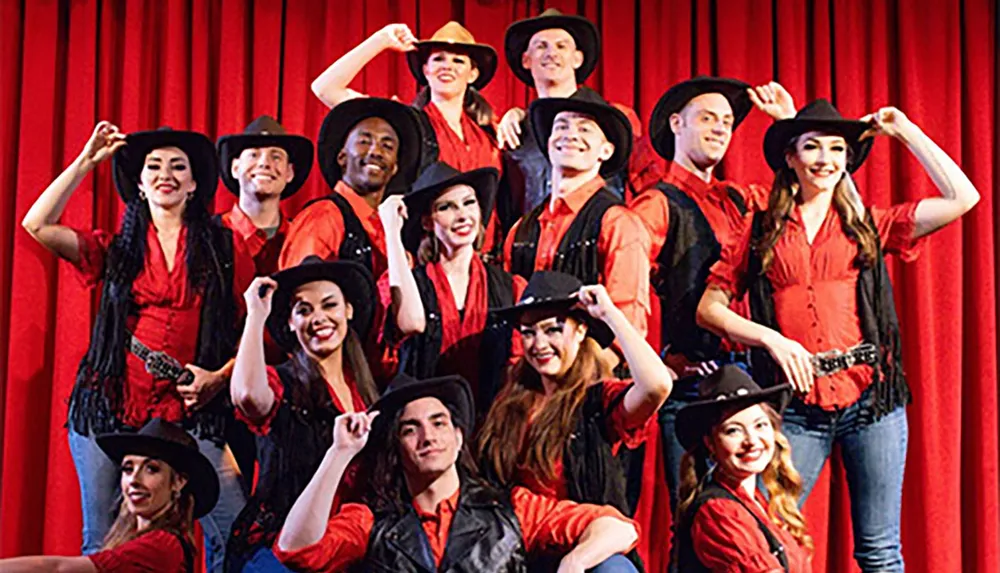 A group of performers dressed in red shirts black hats and country-western attire pose on stage with a red curtain backdrop smiling enthusiastically