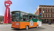 The image shows a colorful hop-on hop-off tour trolley filled with passengers in front of urban scenery, featuring a distinctive red abstract sculpture.