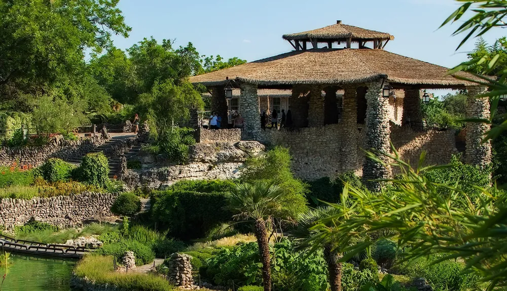 This image features a rustic stone building with a thatched roof surrounded by lush greenery and a small pond with people exploring the serene outdoor setting