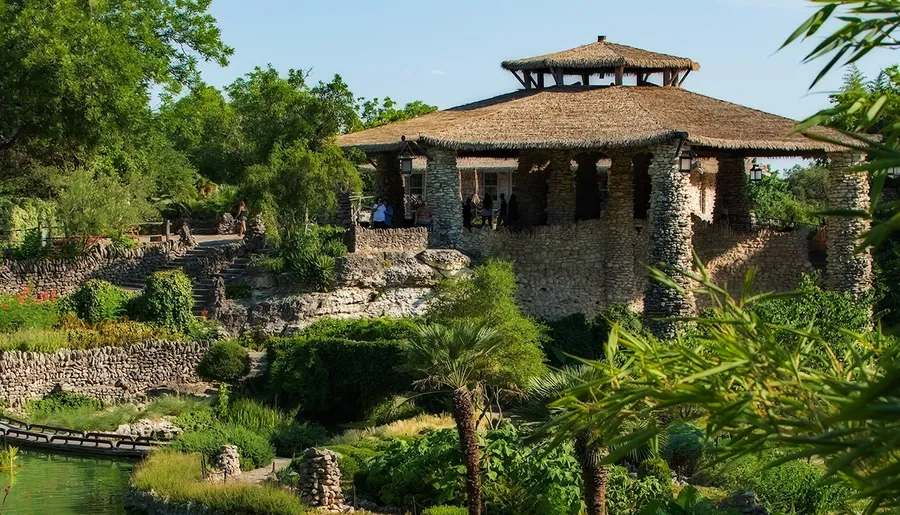 This image features a rustic stone building with a thatched roof, surrounded by lush greenery and a small pond, with people exploring the serene outdoor setting.