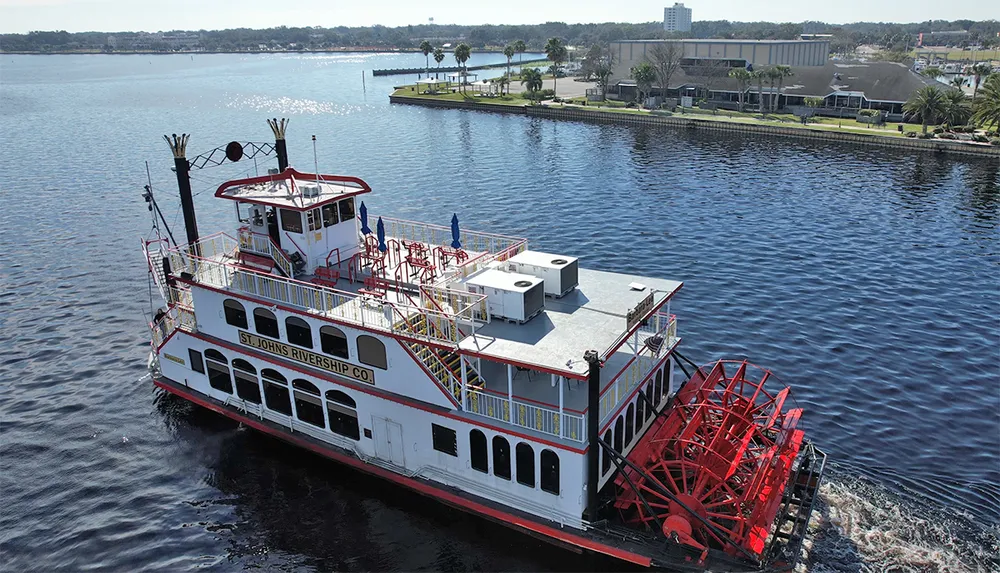 A paddlewheel riverboat is cruising along the calm waters of a river with a shoreline featuring a park and buildings in the background