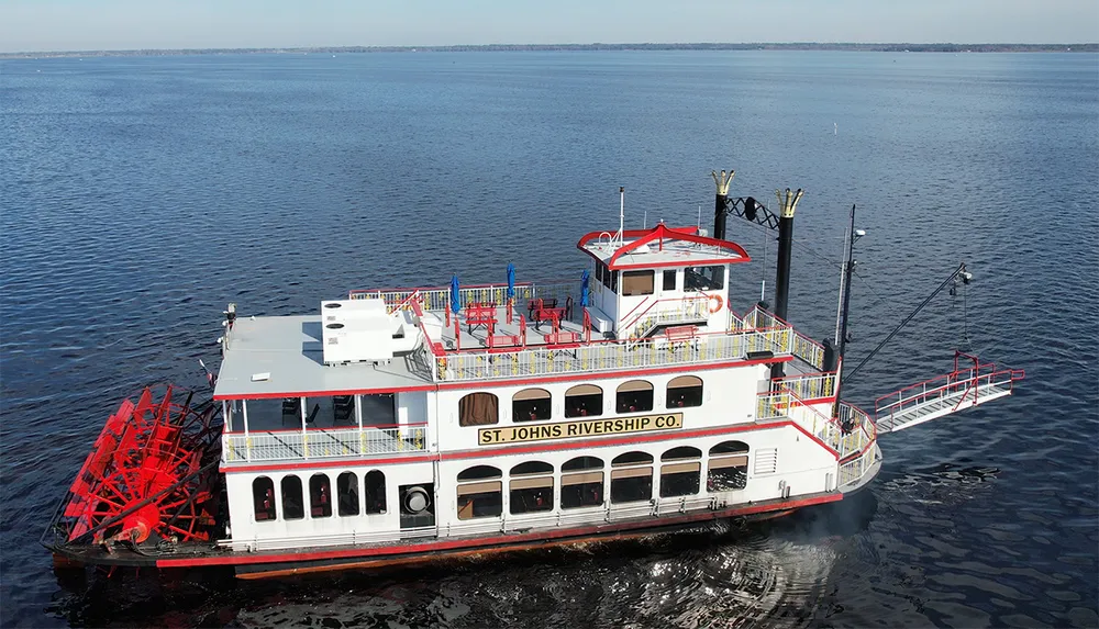 A red and white paddlewheel riverboat is moored on a calm body of water under a clear blue sky