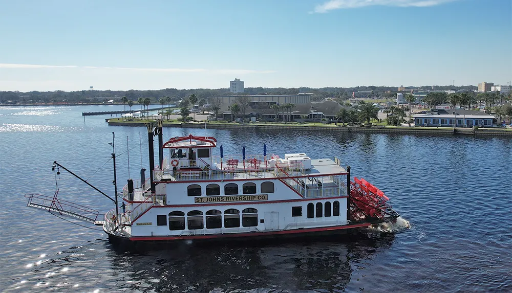 A paddlewheel riverboat from the St Johns Rivership Company is cruising on a calm river with urban buildings and a clear blue sky in the background
