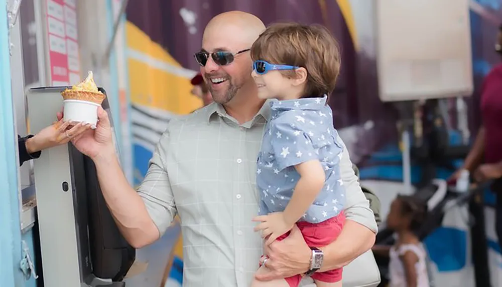 A smiling man carrying a young child is accepting an ice cream cone from a vendor at an outdoor event