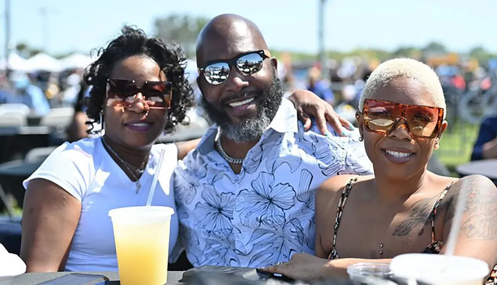 Three smiling people are enjoying an outdoor event together with a man in the center wearing sunglasses and embracing two women on either side of him all in casual summer attire