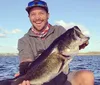 A smiling person proudly holds a large fish with a lake and a clear sky in the background