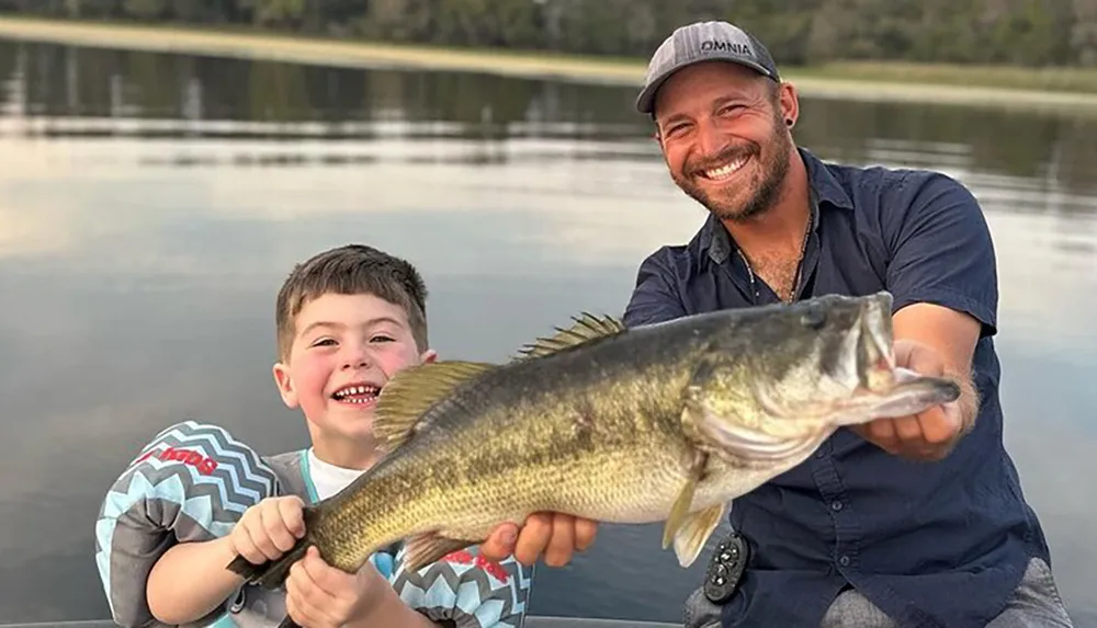 A smiling child and a man are proudly holding up a large fish theyve caught with a serene body of water in the background