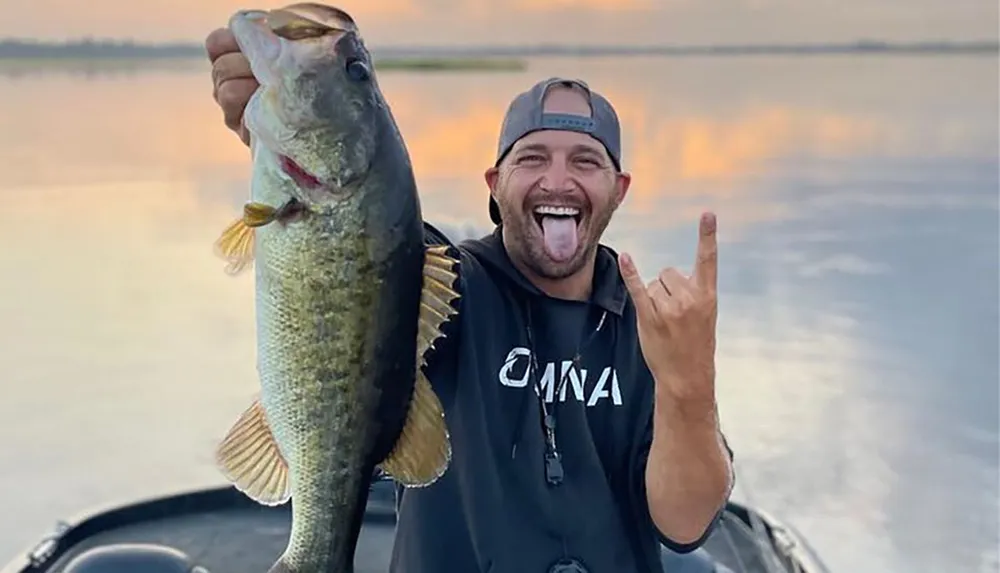 A man is holding a large fish and making a playful face and hand gesture with a water backdrop at sunset