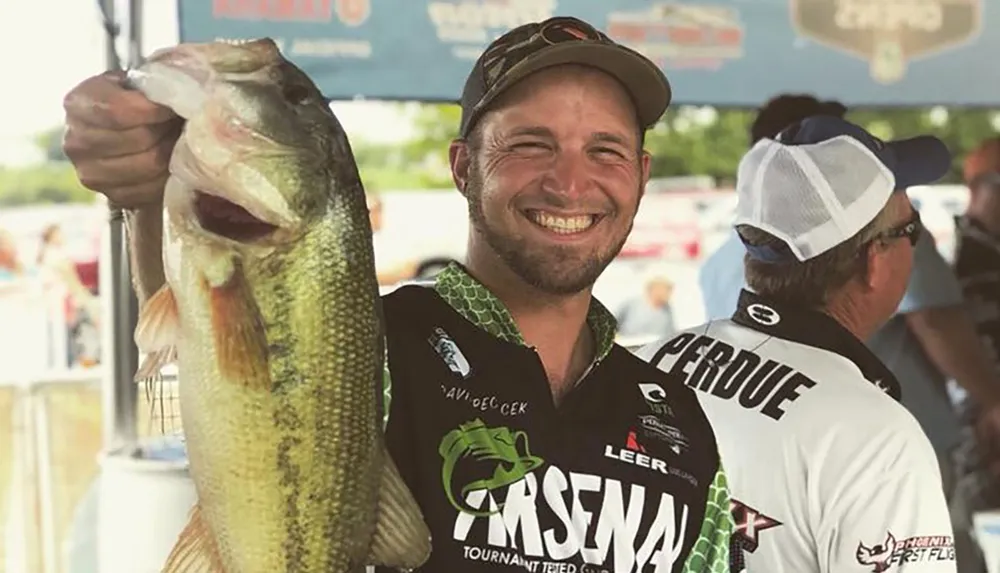 A man is smiling broadly as he holds up a large fish at what appears to be a fishing tournament with event banners in the background and another participant visible