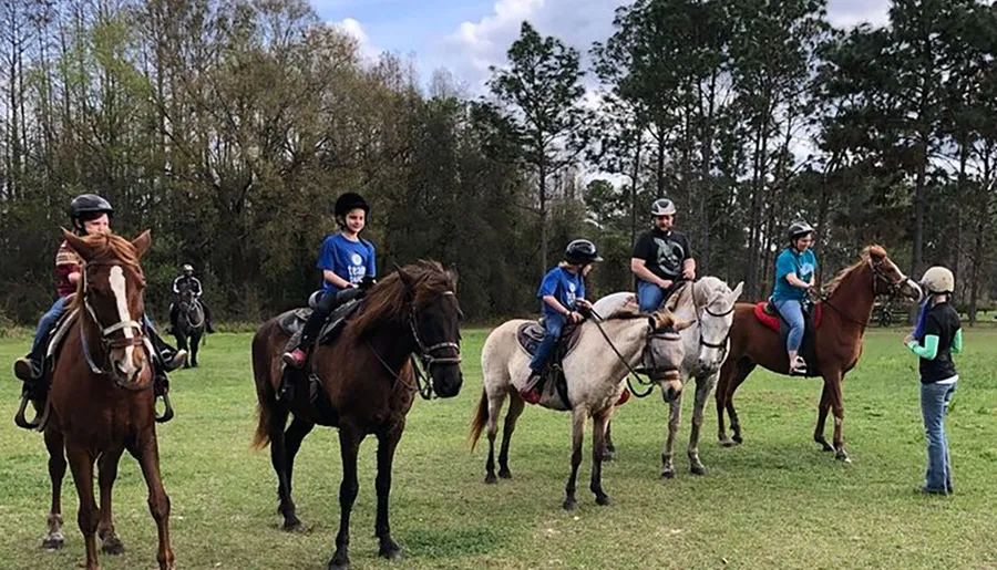 A group of people wearing protective helmets are enjoying a day of horseback riding in a grassy outdoor area with trees in the background.