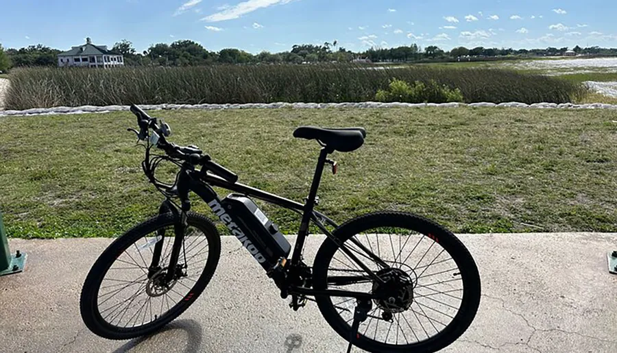 A bicycle is parked on a concrete path with a scenic backdrop of grassland and a house in the distance under a clear sky.