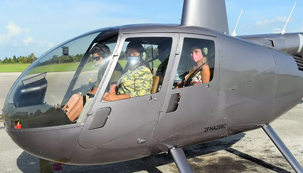 Three people wearing face masks are seated inside the cockpit of a helicopter preparing for a flight
