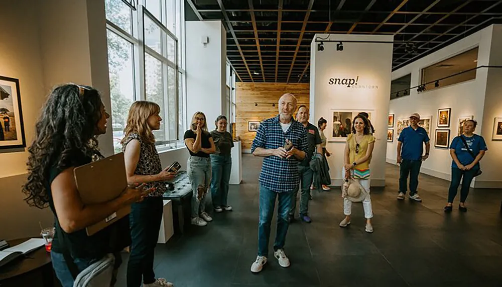 A group of people is attentively listening to a person speaking in an art gallery with photographs displayed on the walls