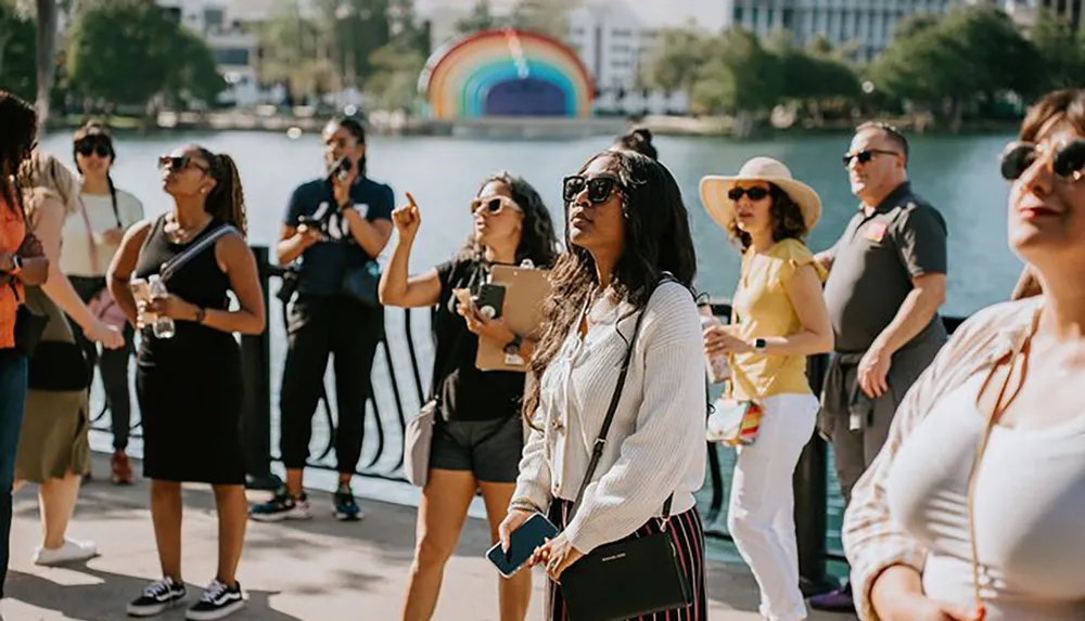A group of people are gathered outdoors by a waterfront seemingly engaged in a guided tour or event with a colorful rainbow arch structure in the background