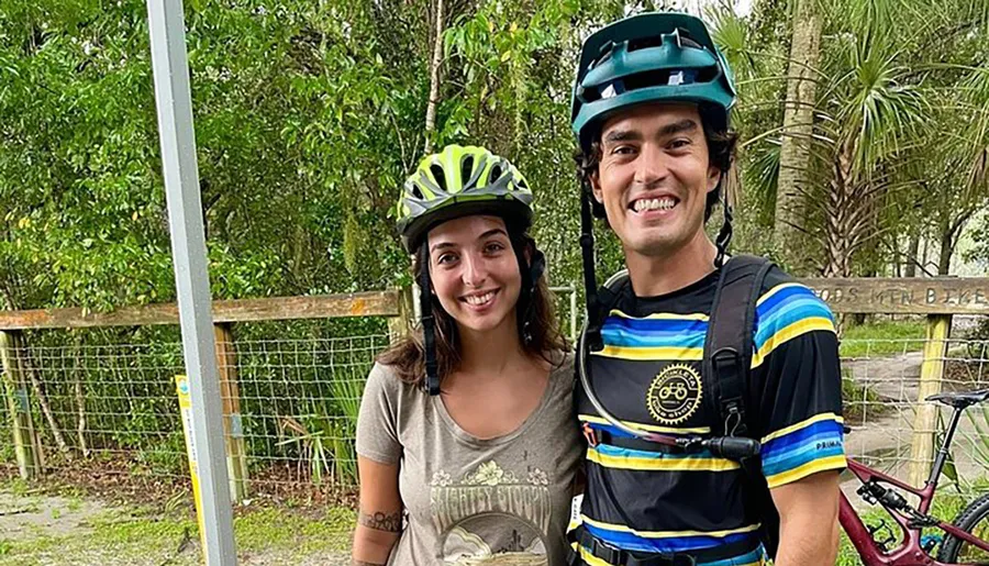 Two happy cyclists wearing helmets are posing for a photo with a bike partially visible in the background.