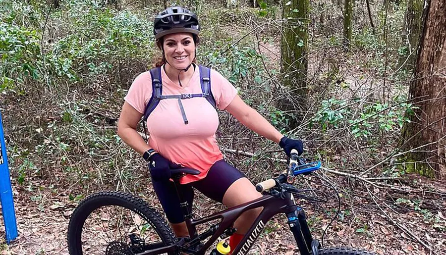 A person is posing with a smile on a mountain bike in a wooded area, equipped with a helmet and hydration pack.