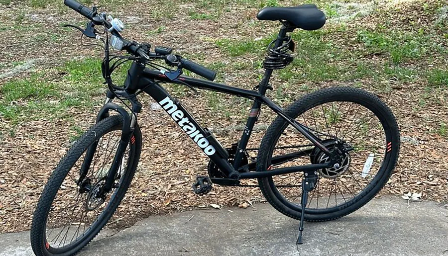 The image shows a black mountain bike with the brand name 'Metakoo' displayed on its frame, stationed outdoors on a concrete surface with grass and fallen leaves in the background.